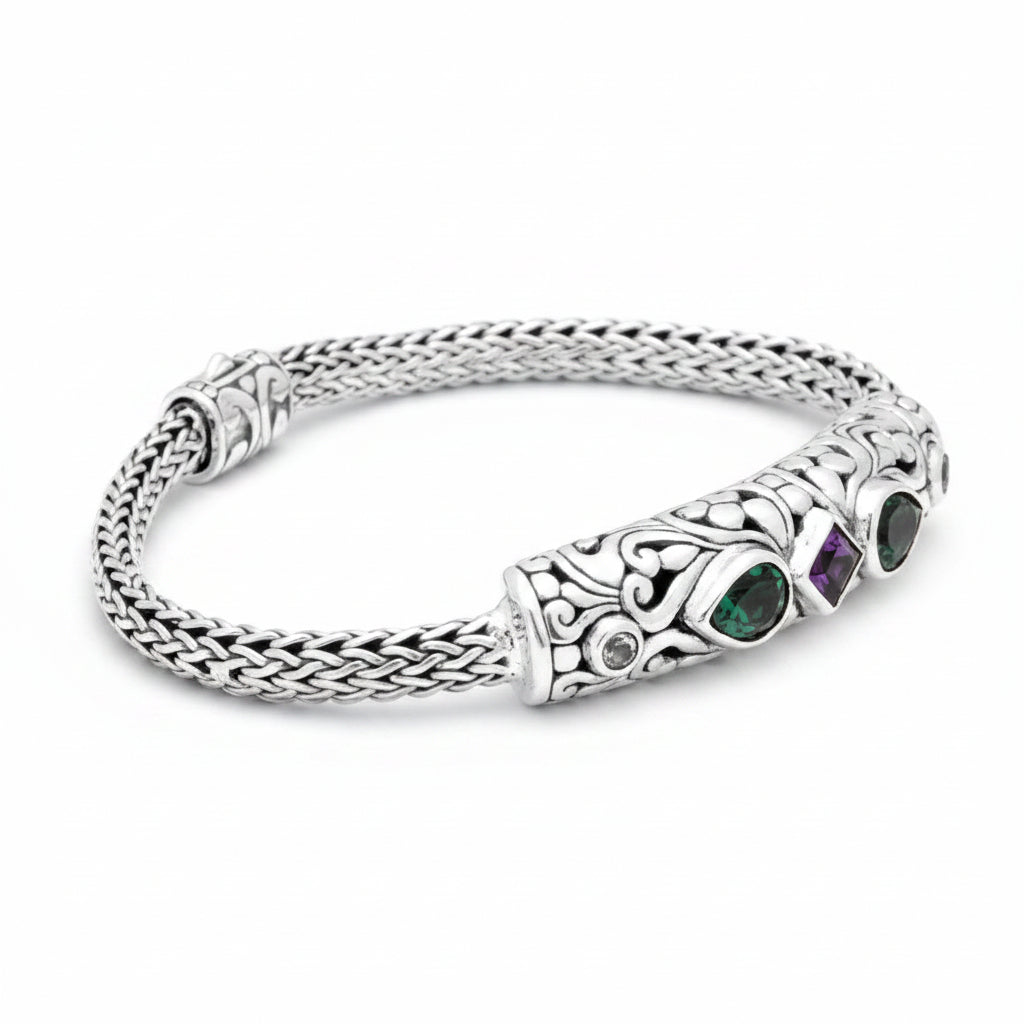 Bracciale Tulang Naga Tre Gemme Balinesia su sfondo bianco, vista dall'alto – catena intrecciata a mano in argento 925 ossidato con barra centrale decorata a volute, smeraldo, ametista e topazio – Collezione Viaggio a Celuk