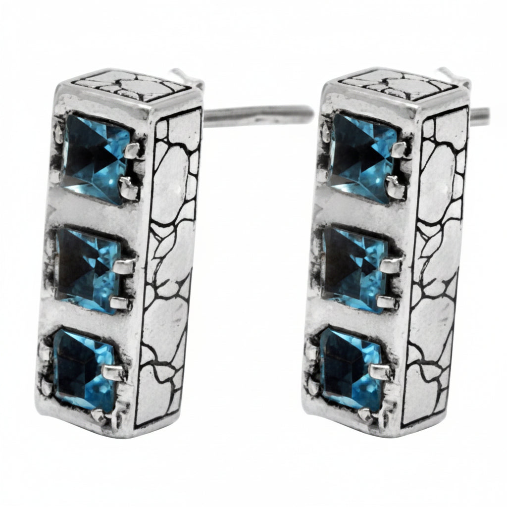 Orecchini Sinar Sky Blue Topaz in argento 925 ossidato, vista 3/4 su sfondo bianco, barra rettangolare 19×7 mm con 3 topazi blu quadrati a griffe e texture Crocodile – Balinesia