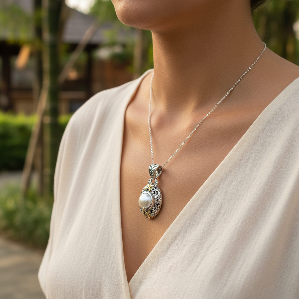 Ciondolo Lovina Balinesia, vista frontale su sfondo bianco con perla Mabé rotonda naturale in castone bezel, due fiorellini in oro 18k e volute traforatein argento 925 ossidato