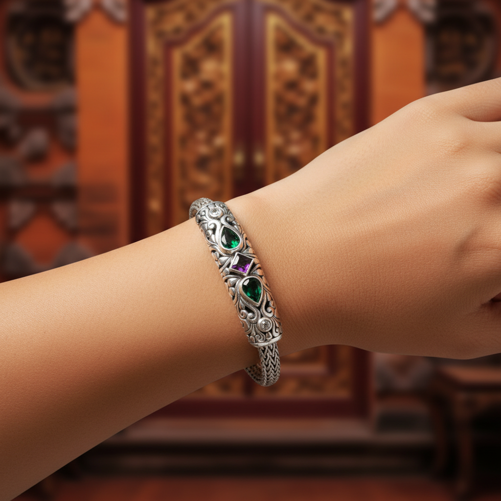 Bracciale Tulang Naga Tre Gemme Balinesia indossato su polso femminile – smeraldi verdi e ametista viola visibili sulla barra decorata in argento 925 ossidato, sfondo porta balinese intagliata – Collezione Viaggio a Celuk