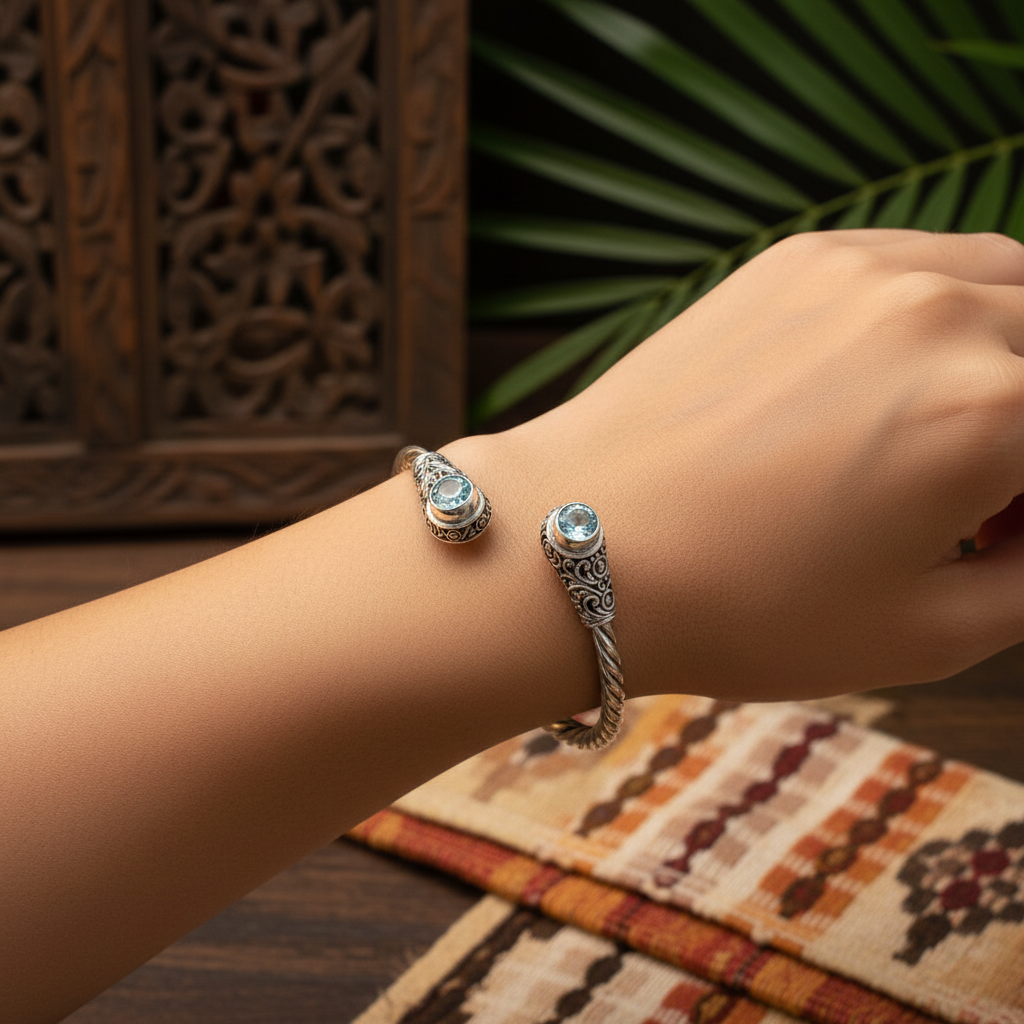 Bracciale Mata Air Balinesia indossato al polso – argento 925 ossidato con torchon e topazi azzurri naturali, sfondo con pannello intagliato balinese e tessuto batik – Collezione Viaggio a Celuk